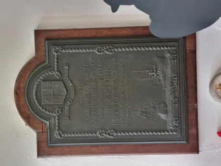 A metal plaque with a wooden backboard with a knotted rope decorative border, family crest with the inscription commemorating the military service of the subject below. With images of HMS Algiers and HMS Exmouth below the inscription.