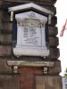 Wall mounted white stone memorial on slate backboard.  Protective pitched wooden "roof" above, stone shelf below