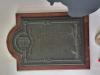 A metal plaque with a wooden backboard with a knotted rope decorative border, family crest with the inscription commemorating the military service of the subject below. With images of HMS Algiers and HMS Exmouth below the inscription.
