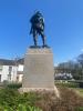 A stone base topped with a statue of a soldier with his back turned. There's an inscription on the stone. It's surrounded by a garden.