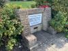 Full view of small brick wall in a park landscape with main memorial plaque above a much smaller brass plaque.