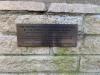 Small rectangular brass plaque with inscription in black lettering against brick background wall.