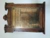 Wall-mounted brass plaque with dark wooden ornate frame. Black incised lettering (in Welsh). 
