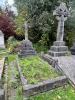 Grey granite memorial at the head of a kerbed grave infilled with grass, consisting of a base of three square plinths topped by a rock motif effect and surmounted by a latin cross adorned with garlands. Inscriptions are written on all three square plinths and the rock motif. The relevant commemoration is on the lowest square plinth. 