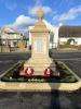 A bright stone memorial stands a the centre of a road junction. Poppy wreaths at its base.