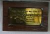 A small brass plaque within a wooden frame attached to the wall just below the main memorial listing the donors