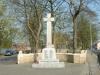 Stone cross on a cuboid plinth with a square base against a low stone wall and trees in a green by a main road. 